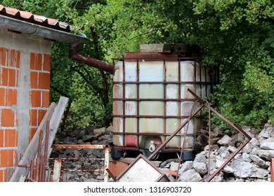 Vintage Old Partially Rusted Metal And Plastic Water Container Connected To Gutter To Collect Rain Water Mounted On Stone Pile Surrounded With Construction Material And Dense Trees On Warm Sunny Day