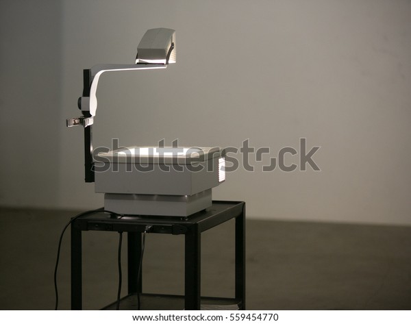 A vintage overhead projector sit on a roller\
cart lighting a wall ready to show overhead projection\
transparencies. Overhead projectors were often used in school\
& business before digital\
projection.