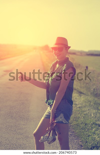 Vintage photo of traveler woman hitchhiking along\
lonely road in summer\
sunset
