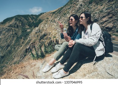 Vintage Picture Two Asian Female Best Friends Hikers Sitting On Rock Mountain Cliff While Holding Mobile Phone Pointing Finger Far Away To Pacific Ocean View. Young Girl Travelers Relaxing On Big Sur