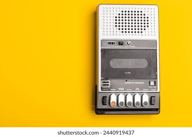 VIntage portable tape recorder with open cover and compact cassette inside compartment on yellow background. The device features built-in speaker microphone and simple control buttons at the bottom