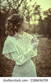 Vintage portrait of a beautiful noble lady in a white dress walking in the old park. Hairstyle and makeup, late 19th century. Historical reconstruction of the Victorian era. 