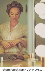 Vintage Portrait Of A Pretty Young Blonde Woman With Curls On Her Head, Getting Dressed In Front Of Her Dressing Table Topped With A Mirror Framed With Light Bulbs