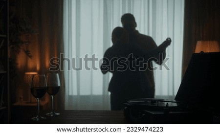 Vintage record player with a vinyl record and glasses with red wine on the table close up. A young couple is dancing a slow dance while enjoying a pleasant evening together. Silhouette.