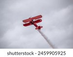 A vintage red biplane flying with a trail of smoke.