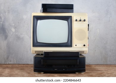 Vintage Retro Analog Small TV Set With VHS Video Player And Videocassetes Stand On Wooden Floor Front Grey Concrete Wall Background.