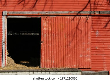 Vintage Retro Bright Red Metal Farm Barn Loading Dock Door Open With Livestock Feed Hay And Tree Shadows As Agricultural Architectural Scene