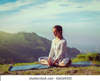 Vintage retro effect hipster style image of sporty fit woman practices yoga asana Baddha Konasana - bound angle pose outdoors in HImalayas mountains in the morning with sky. Himachal Pradesh, India