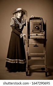 Vintage retro style photo of a young woman in a long modest dress and hat standing near a daguerreotype vintage camera on a gray background. Retro photography and vintage concept.