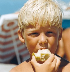 Vintage Roll Film Retro 1979 Image, Close-up Portrait Of A Young Boy With Blond Hair And Beach Umbrella Background Eating A Green Apple.