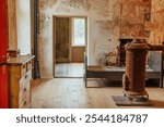 A vintage room featuring an antique iron stove, rustic bed, and worn dresser. The walls show peeling paint, giving a nostalgic charm, illuminated by soft sunlight through the window