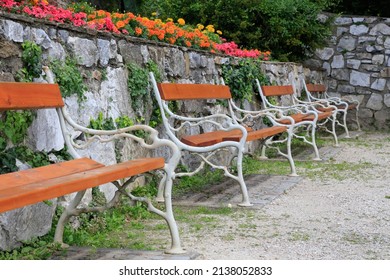 Vintage or rural garden design: Enchanting resting place with a line of wrought iron benches with wooden seats in front of an overgrown quarry stones wall which is decorated with blooming flowers