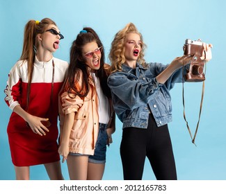 Vintage selfie. Pretty young women in retro 90s fashion style, outfits isolated over blue studio background. Concept of eras comparison, beauty, fashion and youth. Look happy, excited, delighted