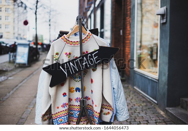 Vintage\
sign with a background of different vintage clothing on a street.\
White vintage sweater with embroidered flowers. \
