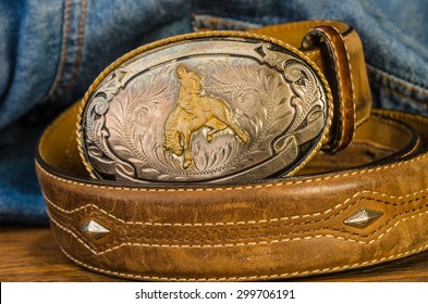 Vintage silver buckle with cowboy on bucking bronc.  Leather belt with studs against blue denim work shirt background.