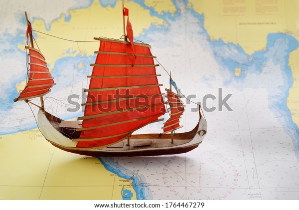 Vintage simple wooden craft\
scale model of a tall ship with red sails and old white nautical\
chart close-up. Planning travel, sailing accessories, concept\
art
