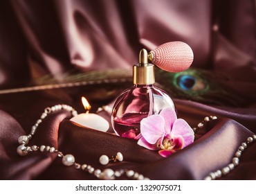 Vintage spray atomizer perfume bottle and pearl jewellery on silky dark pink fabric with peacock feather and candle burning, romantic luxurious evening concept. 