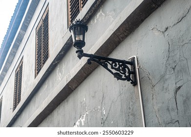 A vintage street lamp with intricate wrought iron design is mounted on an old, weathered building with wooden shutters, highlighting the rustic charm of historical architecture - Powered by Shutterstock