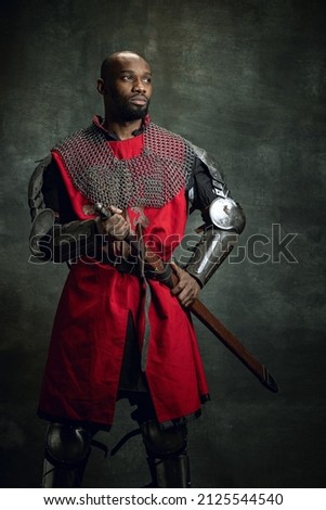 Vintage style portrait of brutal dark skinned man, medieval warrior or knight with wounded face wearing armour isolated over dark background. Comparison of eras, history, renaissance style