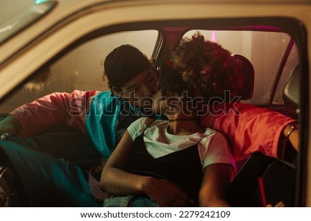 A vintage styled couple is in a car, kissing in the parking lot illuminated by neon lights.