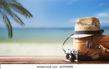 Vintage-Koffer, Hip-Hut, Fotokamera und Reisepass auf Holzdeck. Tropisches Meer, Strand und Palmen drei im Hintergrund. Design-Banner für Sommerurlaubsreisen mit Kopienraum.