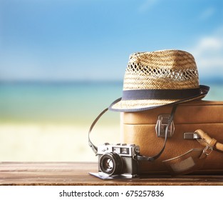 Vintage Suitcase, Hipster Hat, Photo Camera And Passport On Wooden Deck. Tropical Sea And Sandy Beach A In Background. Summer Holiday Traveling Design Concept.