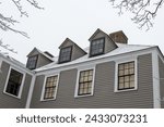 A vintage tan color wooden building with white trim, multiple double hung windows, dormers, gables, a black shingled roof, and a brown brick chimney. The peaked roof is covered in fresh white snow.
