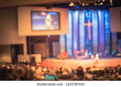 Vintage Tone Blurred Abstract Christian People Inside Church Listening To Preacher Speaking. Defocused Back View Audience On Row Of Raised Sitting Chairs Looking At Stage With Video Projector Screens