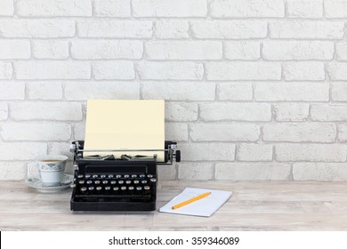 Vintage typewriter, notepad, pencil and china tea cup with tea on old shabby wood effect desk  top with white brick wall background