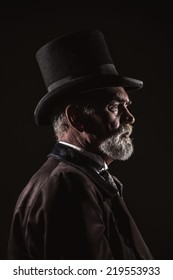 Vintage victorian man with black hat and gray hair and beard. Studio shot against dark background.