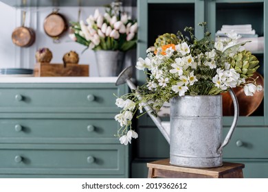 Vintage watering can filled with lots of different flowers and greenery, sitting on top of wooden ladder, comfortable kitchen enviroment, green furniture in blurred background