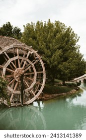 Vintage Watermill Beside a Tranquil River and Stone Bridge. Old wooden waterwheel by a serene lake with an arched bridge