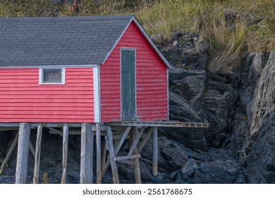 A vintage wooden fishing shed on stilts built into a rocky coastline over water. The fishing stage has a single solid green wood door with a small glass window. The roof is black asphalt shingles. - Powered by Shutterstock