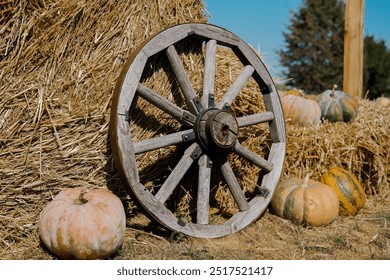 A vintage wooden wagon wheel leans against a stack of hay bales, surrounded by a variety of colorful pumpkins under a clear blue sky, capturing the essence of fall harvest. - Powered by Shutterstock