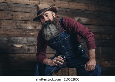 Vintage Worker Man With Long Gray Beard In Jeans Dungarees Holding Whiskey. Sitting On Wooden Crate In Barn.