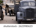 Vintage World War II German military truck on display during a historical reenactment event, parked in front of a war-damaged building set with sandbags and props.