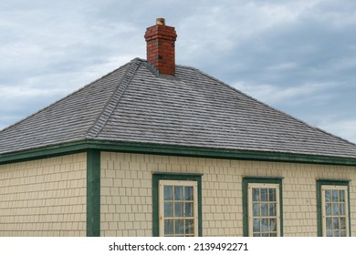 A Vintage Yellow Cedar Shake Clapboard House With A Grey Shingled Wooden Hip Roof. The Building Has A Red Brick Chimney And Multiple Glass Windows.  The Sky In The Background Is Blue With Clouds.
