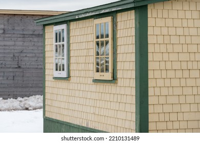 A Vintage Yellow Cedar Shake Clapboard House With A Grey Shingled Wooden Roof. The Building Has Two Glass Storm Windows With Three Small Air Holes.  There's A Grey Building In The Background.