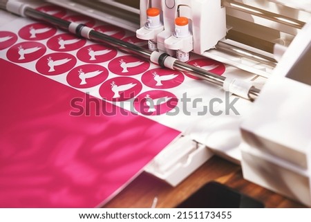 vinyl plotter cuts round stickers with peace dove symbol from pink adhesive foil in vivid shadowy light. view on tool head, guides, excessive foil and wooden worktop. peace movement and DIY concept
