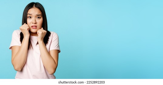 Violance, abuse victim concept. Scared timid asian woman shaking in fear, terrified, looking insecure and frightened, standing over blue background.