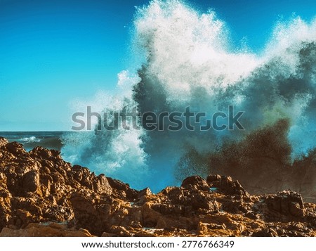 Violent ocean wave explodes dramatically over jagged, reddish-brown rocks, under a clear blue sky