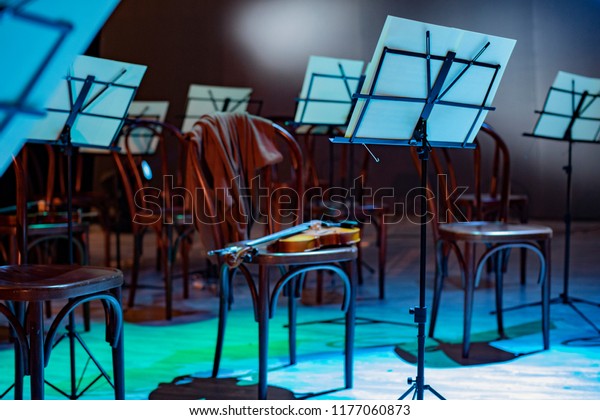 Violin and music stand. Violin on a chair and music\
stands with notes around. the orchestra is on break. sweater on the\
back of a chair
