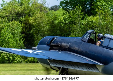 Virginia / USA - May 9 2015: Side Profile Of A Pilot In An Open Cockpit Of A Blue Grumman F4F WW2 Fighter Plane On A Field With Trees In Background