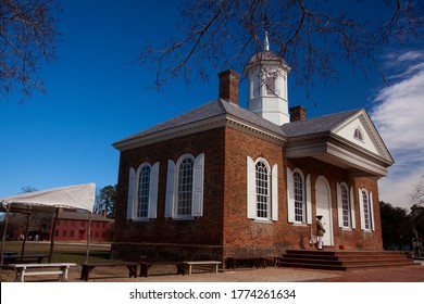 Virginia, USA 02/22/2014: A  Vintage Church With Brick Exterior, Chimneys, Tower And Arched Windows With Wooden Shutters In Colonial Williamsburg. There Is A Minuteman With Tricorn Hat Standing.