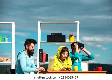 Virtual Business. Modern Technology. Young Men And Women Sitting At A Table With Virtual Reality Goggles. Business Team Testing Virtual Reality Headset In Outdoor Office. Start Up Team Of Freelancers.