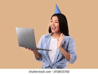 Virtual Celebration Concept. Portrait Of Happy Young Asian Woman Wearing Party Hat Holding Laptop And Waving To Webcam, Making Online Video Call On Computer, Greeting With Birthday, Studio Background