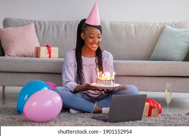 Virtual Celebration. Happy african woman in party hat celebrating birthday online at quarantine or self-isolation, using laptop for video call with friends and family, holding cake, sitting on floor