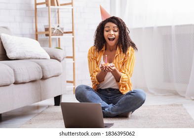 Virtual Celebration. Happy Black Lady In Party Hat Celebrating Birthday Online At Quarantine Or Self-isolation, Using Laptop For Video Call With Friends And Family, Holding Cake, Sitting On The Floor