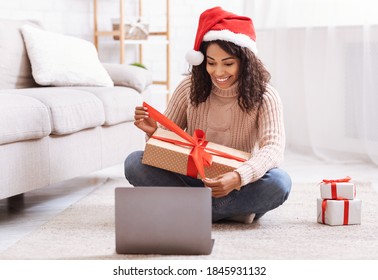 Virtual Celebration. Positive African American Woman In Santa Hat Sitting With Christmas Present On Lap And Opening, Unwrapping Gift Box In Living Room, Sitting On Floor, Making Video Call With Laptop