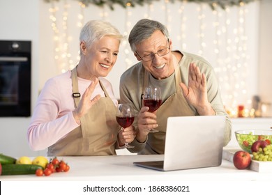 Virtual Christmas Celebration. Happy Senior Couple At Laptop Making Video Call Toasting Celebrating Xmas And Waving Hands Holding Wine Glasses In Kitchen At Home. Distance Christmas And New Year Party